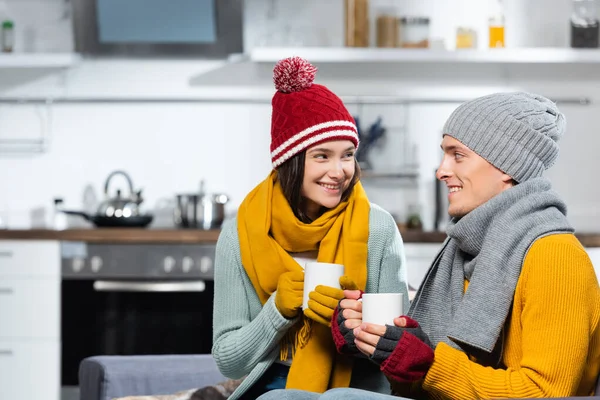 Coppia gioiosa in caldi cappelli, sciarpe e guanti guardarsi mentre tiene in mano tazze di bevanda riscaldante in cucina — Foto stock