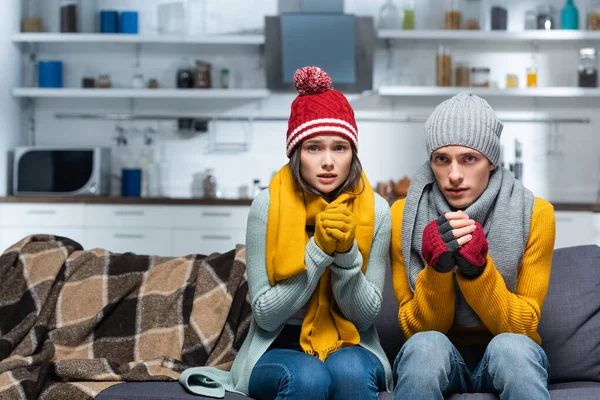 Freezing couple in warm hats and gloves looking at camera while sitting on sofa in cold kitchen — Stock Photo