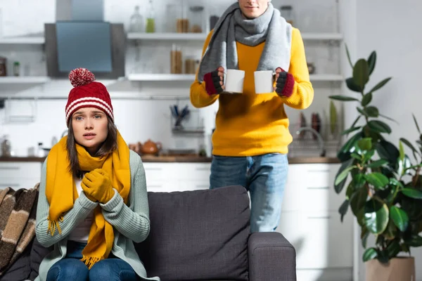 Congelamento donna seduta sul divano in maglia cappello e guanti mentre l'uomo porta tazza con bevanda riscaldante — Foto stock