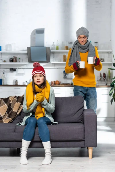 Young man carrying cups of warming drink while freezing girlfriend sitting on sofa in knitted hat and gloves — Stock Photo