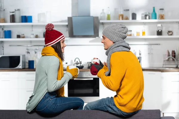 Vista lateral de pareja joven en sombreros calientes, bufandas y guantes sosteniendo tazas con té en la cocina - foto de stock