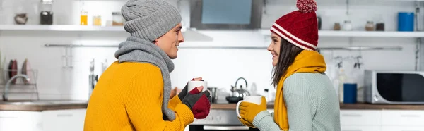 Vista lateral do casal alegre em chapéus de malha, lenços e luvas olhando um para o outro, enquanto segurando copos com chá quente na cozinha, conceito panorâmico — Fotografia de Stock