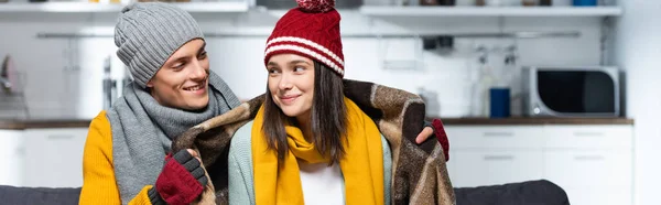 Horizontal image of man in knitted hat wrapping joyful girlfriend with warm plaid blanket while sitting in cold kitchen — Stock Photo