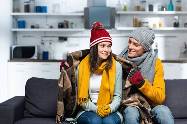 Joven en sombrero caliente y guantes que cubren novia congelante con manta a cuadros mientras está sentado en el sofá en la cocina fría - foto de stock