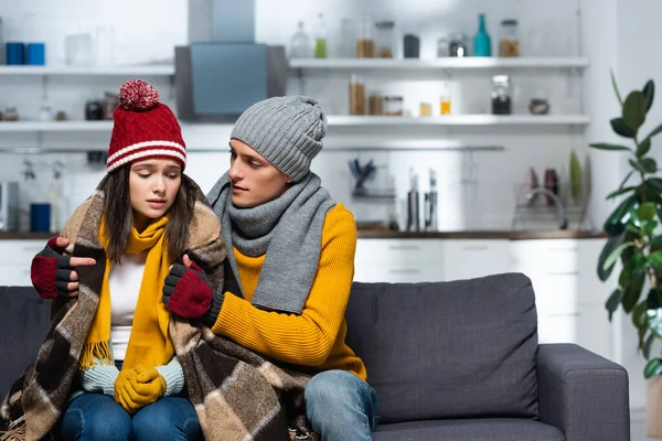 Young man in warm hat and gloves wrapping cold girlfriend with plaid blanket while sitting in kitchen — Stock Photo