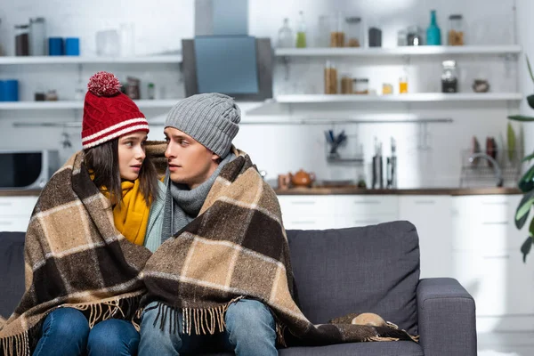 Freezing couple in warm hats looking at each other while sitting under plaid blanket in cold kitchen — Stock Photo