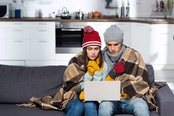 Freezing couple in warm hats and gloves watching movie on laptop while sitting on sofa under plaid blanket — Stock Photo