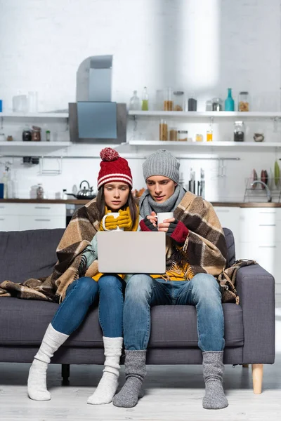 Холодная пара в теплых шляпах и перчатках сидит на диване под клетчатым одеялом и смотрит фильм на ноутбуке — стоковое фото
