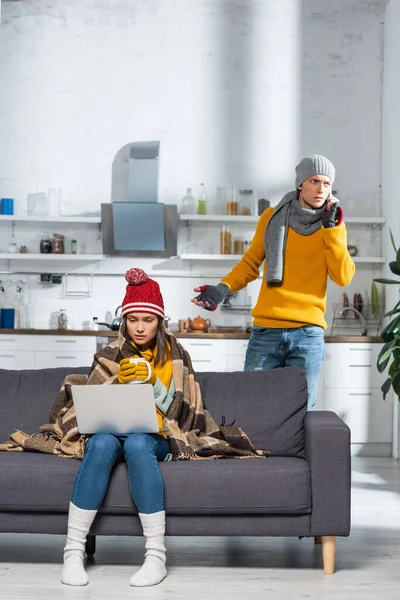 Mujer congelada sentada en el sofá con el ordenador portátil mientras su novio preocupado hablando en el teléfono inteligente en la cocina fría - foto de stock