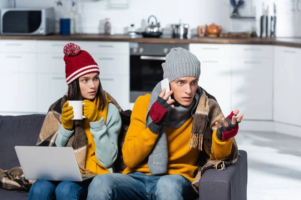 Worried man talking on smartphone near cold woman wearing warm hat nad gloves while sitting with laptop in cold kitchen — Stock Photo