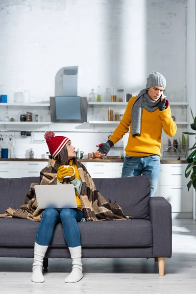 Freezing woman in warm hat and gloves sitting on sofa with laptop while tense boyfriend talking on smartphone — Stock Photo