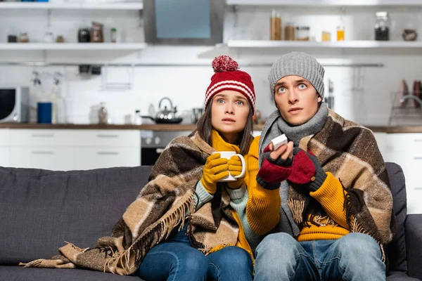 Freezing couple in warm hats, wrapped in plaid blanket, watching tv while sitting in cold kitchen — Stock Photo