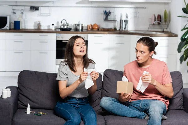 Young man holding paper napkins near sick woman sneezing while sitting near pills and nasal spray — Stock Photo
