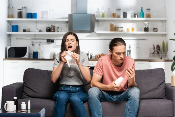 Sick woman sneezing near man holding bottle with medicines while sitting in kitchen — Stock Photo