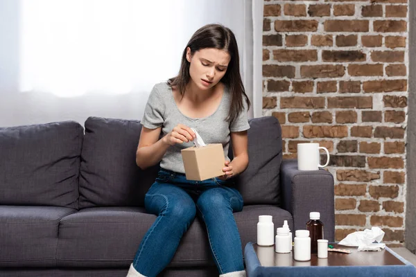 Mujer enferma que toma servilleta de papel mientras está sentada cerca de la taza de bebida caliente y la mesita de noche con medicamentos - foto de stock