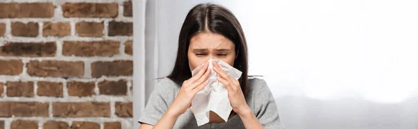 Imagen horizontal de la mujer enferma estornudando en la servilleta de papel mientras está sentada en el sofá en casa - foto de stock