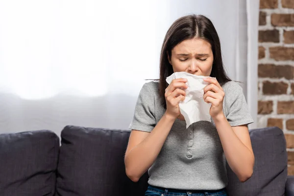 Kranke Frau niest in Papierserviette, während sie zu Hause auf Sofa sitzt — Stockfoto