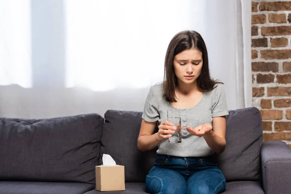 Больная женщина держит стакан воды и таблетки, сидя на диване возле бумажных салфеток — стоковое фото