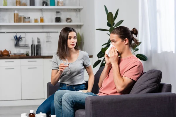 Giovane donna che tiene un bicchiere d'acqua vicino all'uomo starnutisce in un tovagliolo di carta in cucina — Foto stock