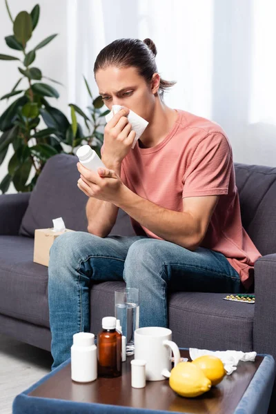 Diseased man wiping nose with paper napkin while sitting on sofa and holding bottle with pills — Stock Photo