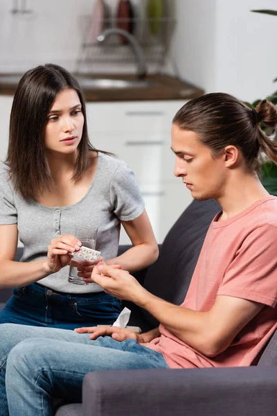 Young woman holding glass of water and giving pills to diseased man sitting on sofa in kitchen — Stock Photo