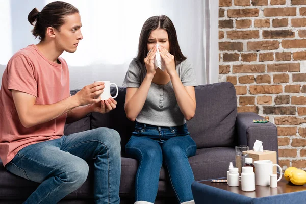 Mann hält Becher mit Warngetränk in der Nähe Frau niest in Papierserviette in der Nähe von Medikamenten — Stockfoto