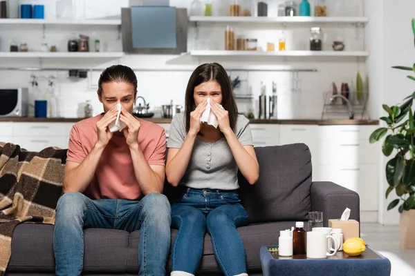 Hombre y mujer enfermos estornudando en servilletas de papel mientras están sentados en la cocina cerca de medicinas - foto de stock