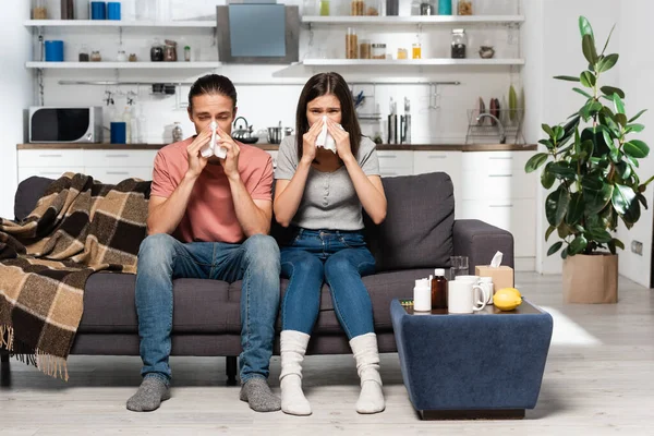 Diseased couple sneezing in paper napkins while sitting on sofa in kitchen near medicines — Stock Photo