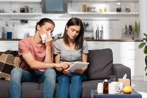 Jeune femme utilisant tablette numérique près malade homme essuyant nez avec serviette en papier et pointant du doigt — Photo de stock