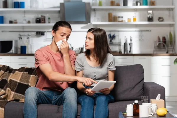 Woman using digital tablet near diseased man pointing with finger and wiping nose with paper napkin — Stock Photo