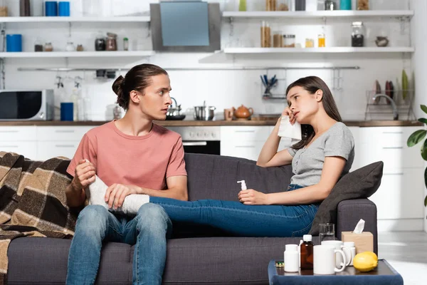 Jeune homme toucher les pieds de la petite amie malade assis sur le canapé avec pulvérisation de gorge — Photo de stock