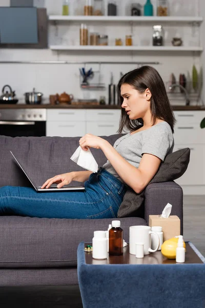Side view of sick freelancer holding paper napkin while sitting on sofa near bedside table with medicines and using laptop — Stock Photo