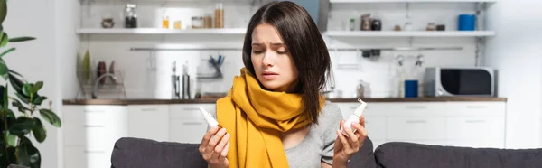 Cultivo panorámico de mujer con bufanda en el cuello sosteniendo aerosoles nasales y de garganta en la cocina - foto de stock