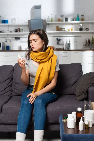 Mulher doente com cachecol no pescoço segurando spray de garganta enquanto sentado perto da mesa de cabeceira com medicamentos — Fotografia de Stock