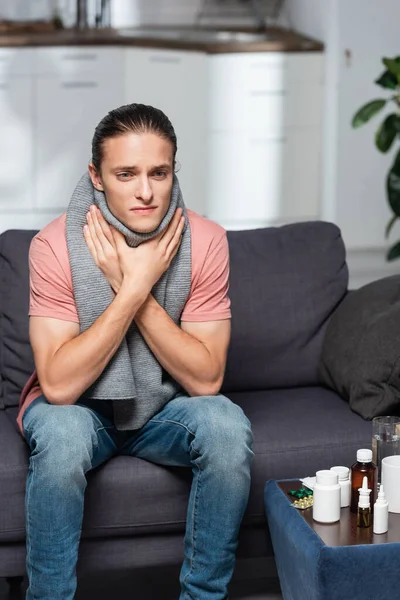 Kranker junger Mann in warmem Schal berührt Halsschmerzen im Sitzen neben Medikamenten — Stockfoto