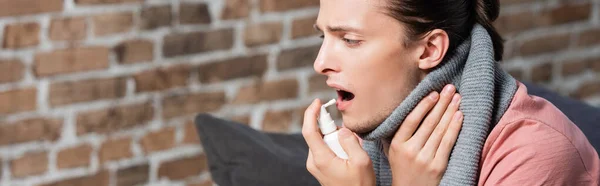 Site web tête de jeune homme malade dans l'écharpe chaude toucher le cou tout en utilisant spray gorge — Photo de stock