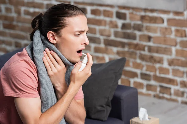 Malato giovane uomo in sciarpa calda utilizzando spray gola mentre toccare il collo — Foto stock