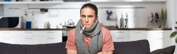 Horizontal image of diseased young man with warm scarf on neck and thermometer in mouth looking at camera while sitting in kitchen — Stock Photo