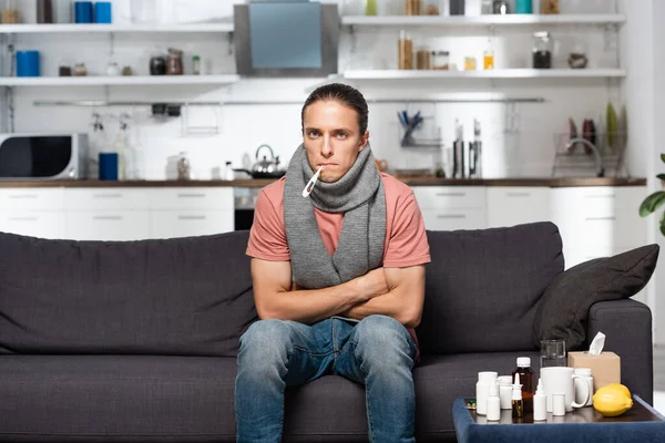 Ill man with warm scarf on neck and thermometer in mouth sitting with crossed arms near medicines — Stock Photo