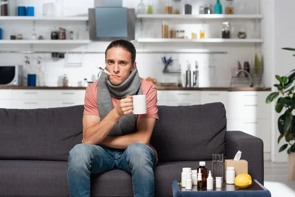 Hombre joven enfermo con termómetro en la boca sosteniendo la taza de bebida caliente mientras está sentado cerca de medicamentos en la cocina - foto de stock