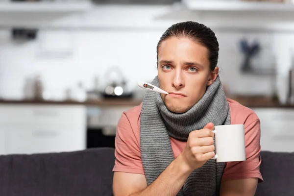 Хворий молодий чоловік з теплим шарфом на шиї і термометром у роті тримає чашку теплого чаю, дивлячись на камеру — стокове фото