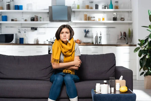 Kranke Frau mit Thermometer im Mund sitzt auf Sofa in Küche neben Medikamenten — Stockfoto