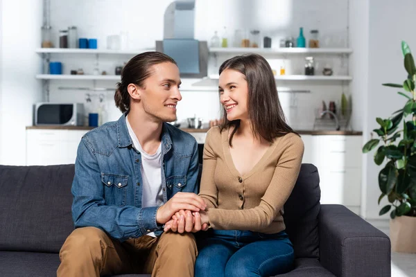 Joven tocando las manos de la novia complacida mientras se sienta en el sofá en la cocina juntos - foto de stock