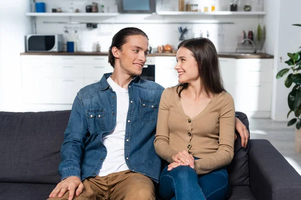 Pareja joven en ropa casual sentada en el sofá en la cocina y mirándose - foto de stock