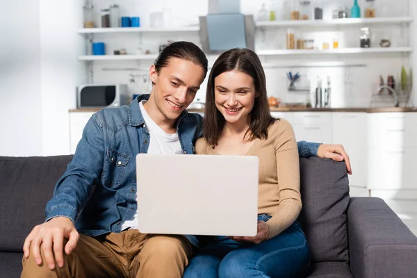 Joyful couple sitting on sofa in kitchen and using watching movie on laptop — Stock Photo