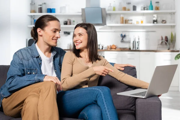 Man sitting near excited girlfriend pointing with finger at laptop on couch in kitchen — Stock Photo