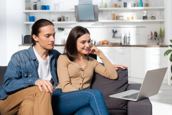 Jeune homme et femme excitée regardant ordinateur portable tout en étant assis sur le canapé dans la cuisine — Photo de stock