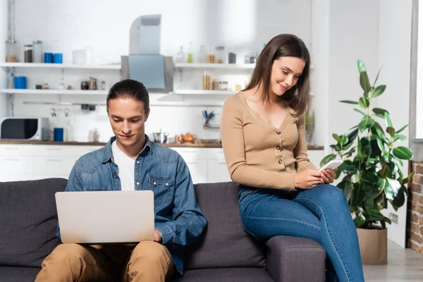 Young man using laptop and woman chatting on smartphone on sofa in kitchen — Stock Photo