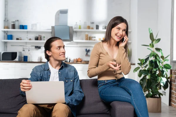 Aufgeregte Frau spricht auf Smartphone neben Mann mit Laptop auf Sofa in Küche — Stockfoto