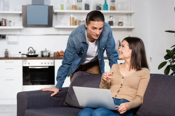Uomo guardando fidanzata eccitato che punta con il dito al computer portatile sul divano in cucina — Foto stock